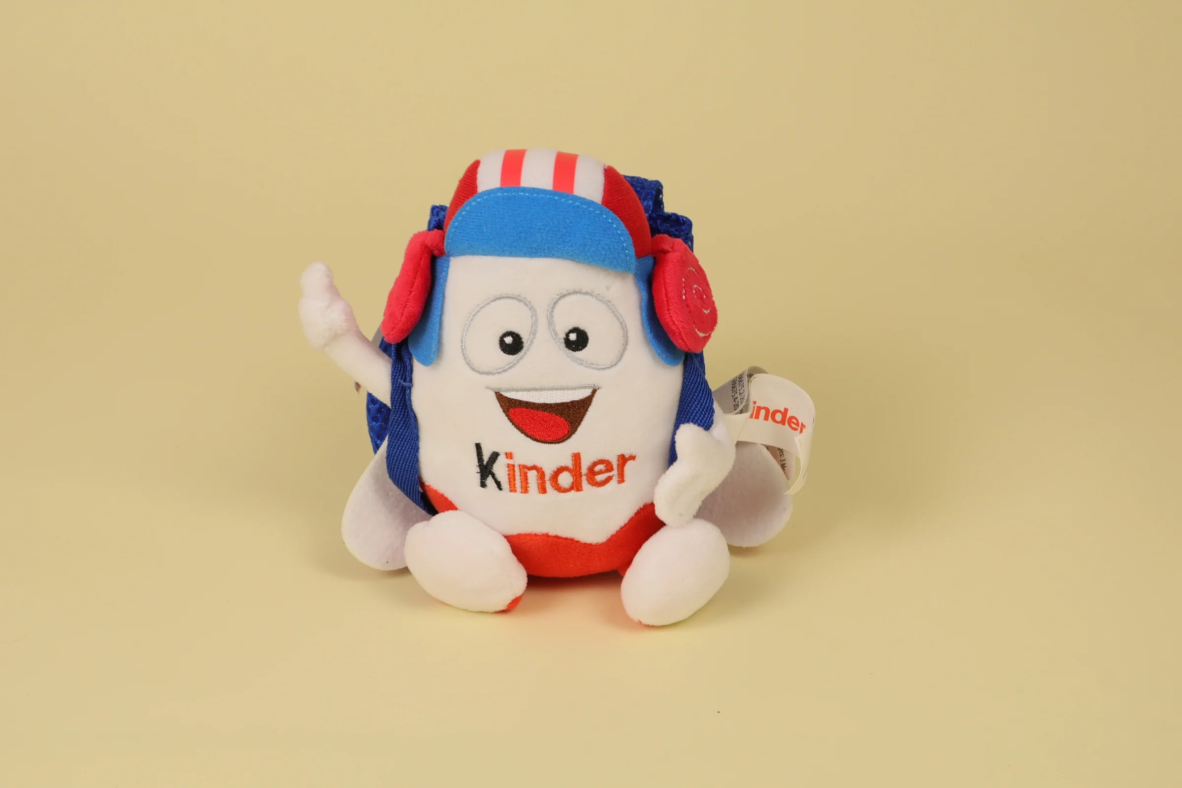 Peluche œuf pilote Kinder de 20 cm avec un casque rouge et blanc et un sac bleu avec des ailes d'avion de la marque Kinder